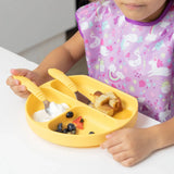 In a purple bib, toddler uses Bumkins Spoon + Fork: Pineapple with a yellow sectioned plate of yogurt, bread, and berries at the table.