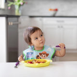 A toddler in a bib uses Bumkins purple Spoon + Fork to eat fruit from a yellow plate at the kitchen table.