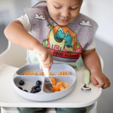 A toddler self-feeds using Bumkins Spoon + Fork: Sage, with fruit and waffles on a sectioned plate, wearing a dinosaur bib.