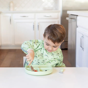 A child in pajamas self-feeds with Bumkins Spoon + Fork: Sage from a food-safe silicone divided plate in the kitchen.