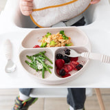 A toddler in a high chair uses Bumkins Spoon + Fork: Sand for green beans, berries, and rice with broccoli on a silicone divided plate.