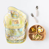 Three colorful bibs with camping designs next to a Bumkins Spoon + Fork: Sand set, perfect for self-feeding adventures.