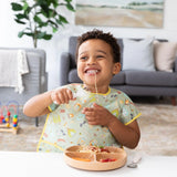 A child in a bib, joyfully self-feeding spaghetti with Bumkins Spoon + Fork: Sand, enjoys toys by a cozy living room sofa.