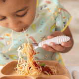 A child self-feeds spaghetti with a Bumkins Sand fork, wearing a patterned bib, using a food-safe beige silicone plate.