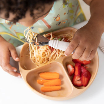 A child self-feeds spaghetti from a silicone plate using Bumkins Spoon + Fork: Sand, with strawberries and carrots included.