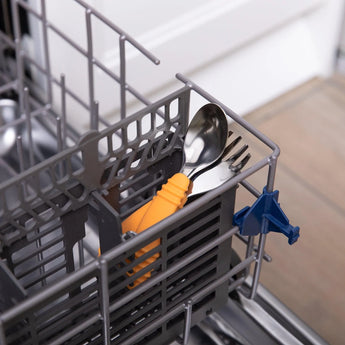 Close-up of a dishwasher cutlery basket with Bumkins Spoon + Fork: Tangerine and an orange-handled toddler utensil inside.