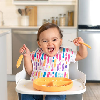 A happy toddler with a colorful bib sits in a high chair, using Bumkins Spoon + Fork: Tangerine over a divided plate of food.