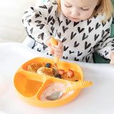 A toddler eats pancakes, fruit, and yogurt from a sectioned yellow plate using Bumkins Spoon + Fork in Tangerine, wearing a heart-patterned shirt.