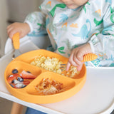 A child uses Bumkins Spoon + Fork in Tangerine to eat scrambled eggs and fruit from a dinosaur-patterned bib on an orange sectioned plate.