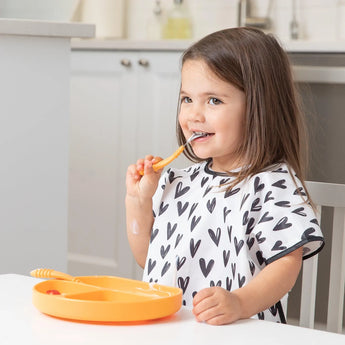 A girl in a heart-patterned shirt uses Bumkins Spoon + Fork: Tangerine to eat from a matching plate at the white kitchen table.