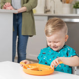 A child in a blue shirt uses Bumkins Spoon + Fork: Tangerine while smiling at an orange plate with an adult nearby in the kitchen.