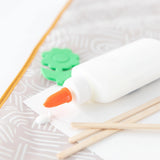 White glue bottle with orange tip, green gear shape, wooden sticks on a Bumkins Splat Mat: Wander background.