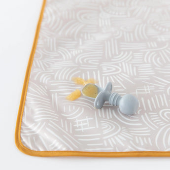 A baby food feeder with soft food pieces sits on the Bumkins Splat Mat: Wander, highlighted by an orange trim design.