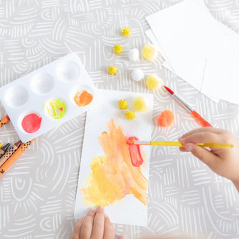 A child paints with orange and pink on paper atop the Bumkins Splat Mat: Wander, surrounded by brushes, a palette, and pom-poms.