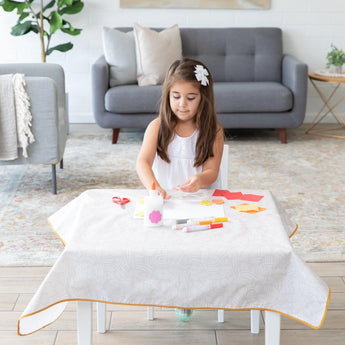 Young girl with a white flower hair clip crafts on a Bumkins Splat Mat: Wander, surrounded by art supplies in a cozy living room.