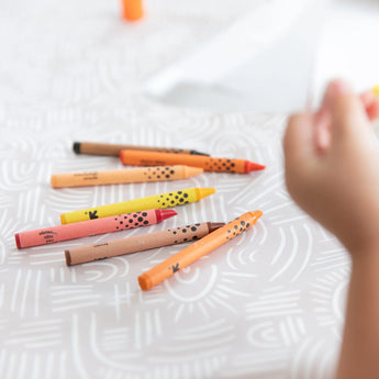 A childs hand reaches for scattered crayons on a white surface, protected by the Bumkins Splat Mat: Wander.