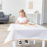 Young girl gluing paper on a Bumkins Splat Mat: Wander, surrounded by craft supplies in a bright living room.