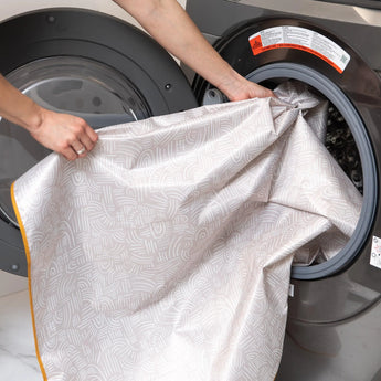 Person loading Bumkins Splat Mat: Wander, a machine washable sheet, into a front-loading washing machine.