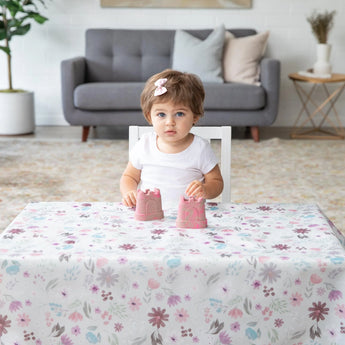 A toddler with short brown hair and a pink bow plays with two small pink sandcastle toys at a table on Bumkins Splat Mat: Floral. In the background, easy cleaning is ensured with the gray sofa, cushions, plant, and decorative table item.