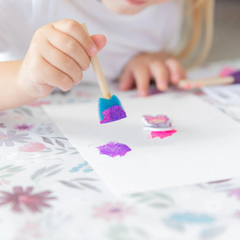 A child paints with a blue and pink sponge brush, using the Bumkins Splat Mat: Floral for easy cleanup.