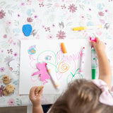 A child draws with colorful pens, nibbling a cookie surrounded by crayons and erasers on a Bumkins Splat Mat: Floral.