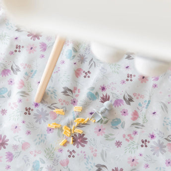 The Bumkins Splat Mat: Floral showcases a broken yellow crayon, sharpener, and a wooden tool on its waterproof design.