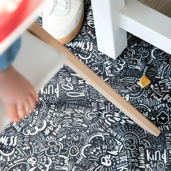 A childs foot and shoe rest near Bumkins Splat Mat: Be Kind, featuring doodles, positive words, and a small orange object.