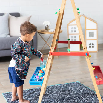 A child paints on a wooden easel at home in a patterned smock, with toys around and a dollhouse, protected by Bumkins Splat Mat: Be Kind.