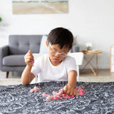 A child with glasses joyfully plays with colorful sand on the Bumkins Be Kind Splat Mat in a cozy living room.