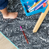 A childs foot on Be Kind Splat Mat by Bumkins, with a blue container of colored pencils, creates a bright scene.