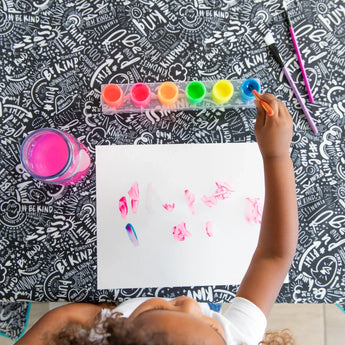 A child paints with colorful paints on white paper at a table covered with the Bumkins Be Kind Splat Mat.