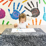 A child colors at a table covered with the Bumkins Splat Mat: Be Kind. Handprint art decorates the wall behind.