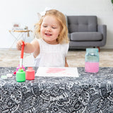 A young girl smiles while painting with bright colors on a Bumkins Be Kind splat mat, with a sofa and small table in the background.