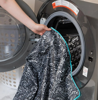 Someone loads the Bumkins Splat Mat: Be Kind, made from waterproof fabric, into a front-loading washing machine.