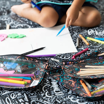 A child draws on paper, sitting on the Be Kind Splat Mat by Bumkins, surrounded by colorful markers and pencils.