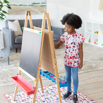 A child in a Minnie Mouse apron paints on an easel with easy-wipe art supplies on Bumkins Splat Mat: Minnie Mouse.