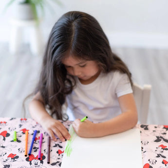 A girl draws with markers on paper at a table covered by the Bumkins Splat Mat: Minnie Mouse.