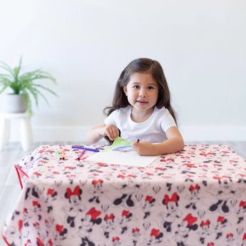 A child draws with markers on a Bumkins Minnie Mouse Splat Mat at a table; a potted plant sits in the background.