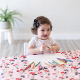 A toddler in a black bow draws at a table covered with a colorful Bumkins Minnie Mouse Splat Mat. A potted plant is behind her.