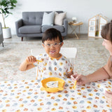 A child with glasses smiles at a table on a Bumkins Splat Mat: Winnie and Friends, holding an adults hand; sofa and plant in the background.