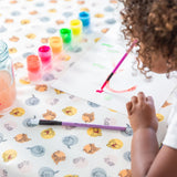 A child paints with watercolors at a table, colorful jars and a water jar on Bumkins Splat Mat: Winnie and Friends for easy cleaning.