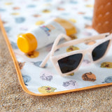 Sunglasses and sunscreen rest on a Bumkins Splat Mat: Winnie and Friends on sandy ground, perfect for easy cleaning after fun in the sun.