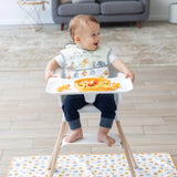 A happy baby in a high chair plays with pasta on a tray while Bumkins Winnie and Friends Splat Mat ensures easy cleanup in the living room.