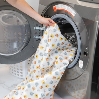 A person loads a washer with Bumkins Splat Mat: Winnie and Friends laundry bag, waterproof, with a basket in the background.