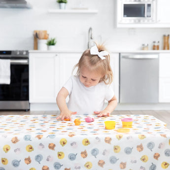 Toddler with a white bow plays on the Bumkins Splat Mat: Winnie and Friends with colorful clay in a bright kitchen.