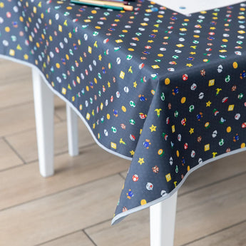 The Splat Mat: Super Mario™ Lineup by Bumkins is a black mat with colorful icons like stars, mushrooms, and coins. Made of waterproof fabric for effortless cleaning, it covers a white table on a light wooden floor. Pencils and paper are scattered across its surface.