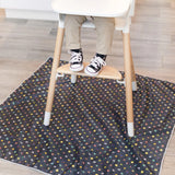 A toddler sits in a high chair on a machine washable Bumkins Super Mario™ Lineup splat mat, wearing beige pants and black sneakers.