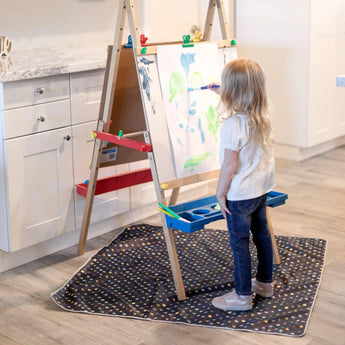 In the kitchen, a child paints on an easel, standing on a Bumkins Super Mario™ Lineup Splat Mat with paints and brushes in trays.