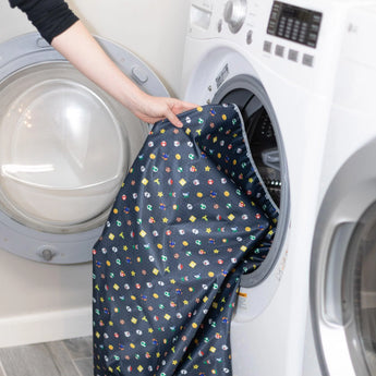 A person places the Bumkins Splat Mat: Super Mario™ Lineup into a front-loading washing machine in the laundry room.