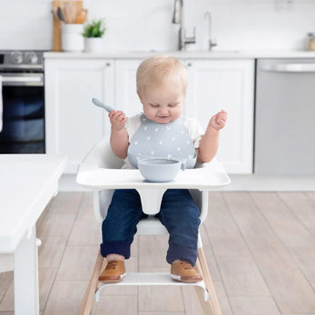 In a bright kitchen, a baby sits in a high chair with Bumkins Silicone Bib: Hearts, holding a spoon and eyeing the bowl on the tray.